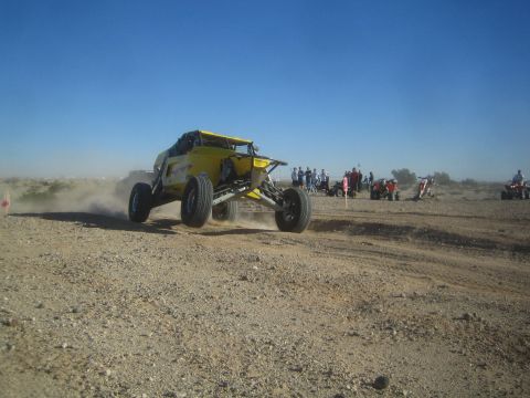 1990 Ford Bronco - yellow tube-chassis off-road buggy goes light over a rise on a desert course as a crowd watches from the ridge behind.