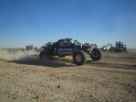 1990 Ford Bronco - black and blue tube-chassis buggy lettered Affordable Raingutters powers through the dust on a desert race course.