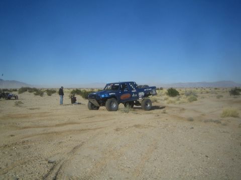 1990 Ford Bronco - Photograph of a blue off-road race truck stopped in the open desert with a person and pit equipment beside it.