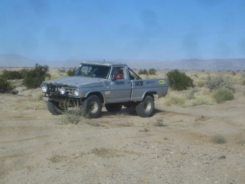 1990 Ford Bronco - Photograph of a silver vintage Ford off-road race truck, number 1478, on a desert trail among brush.