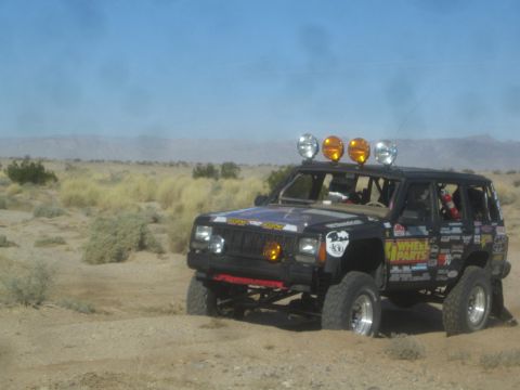1990 Ford Bronco - Photograph of a black sticker-covered Jeep Cherokee race rig with roof light bar driving across the desert.