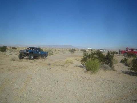 1990 Ford Bronco - Photograph of a blue off-road race truck speeding across the desert with chase trucks visible in the background.