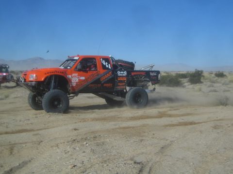 1990 Ford Bronco - Photograph of an orange sponsor-covered off-road race truck, number 1464, charging through the desert.