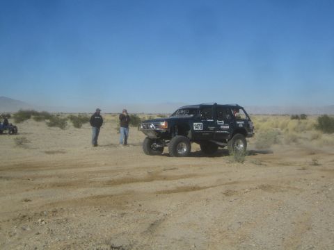 1990 Ford Bronco - Photograph of a dark Toyota off-road race rig on a desert course with spectators standing nearby.