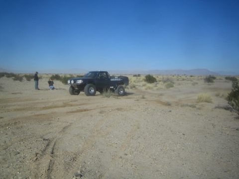 1990 Ford Bronco - Photograph of a black off-road race truck on a dusty desert trail with a few people watching.