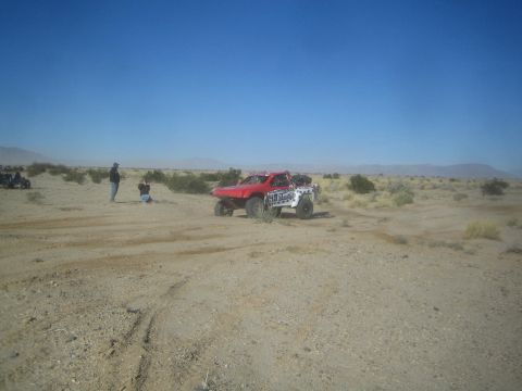 1990 Ford Bronco - Photograph of a red off-road race truck driving across the open desert with onlookers at the side.