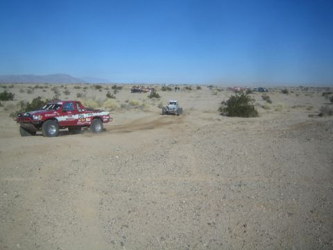 1990 Ford Bronco - Photograph of a red off-road race truck and a buggy navigating a rutted desert course with spectators ahead.