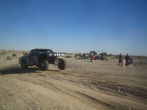 1990 Ford Bronco - Photograph of an off-road race truck with a roof light bar passing a crowd of spectators in the desert.