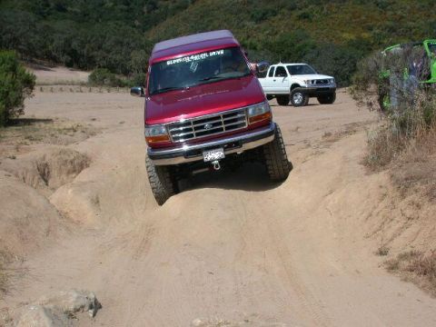 1996 Ford Bronco - A red Ford truck navigates a challenging off-road trail with a white pickup truck in the background.