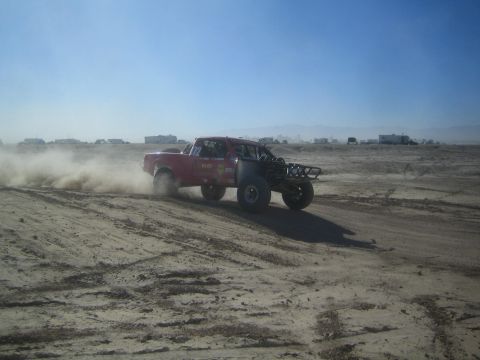 1990 Ford Bronco - Photograph of a red off-road race truck powering through sand and throwing a large dust cloud near a pit area.