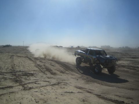 1990 Ford Bronco - Photograph of an off-road race truck cresting a sandy berm with other race vehicles trailing in the dust.