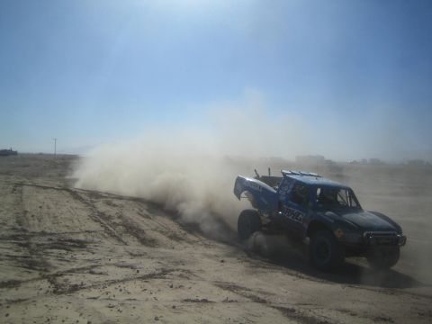 1990 Ford Bronco - Photograph of a blue off-road race truck powering through the desert and throwing a large rooster tail of dust.