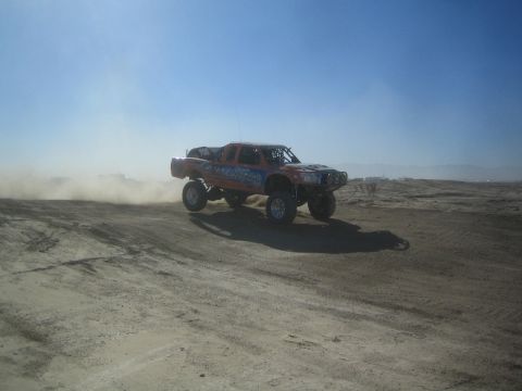 1990 Ford Bronco - Photograph of an orange and blue off-road race truck going airborne over a rise on a dusty desert course.