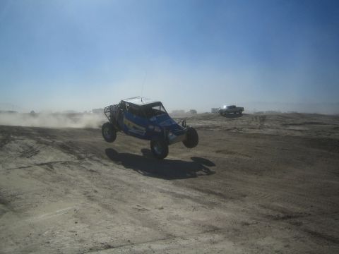 1990 Ford Bronco - Action photo of a blue tube-frame off-road buggy airborne on a dirt course, not a technical document.