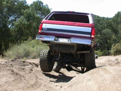 1996 Ford Bronco - Rear view of a red Ford Bronco climbing a dirt berm, with the driver-side rear tire lifted clear of the ground to show the rear axle, drive…