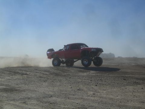 1990 Ford Bronco - Photograph of a red off-road race truck jumping a dirt berm amid blowing desert dust, not a technical document.
