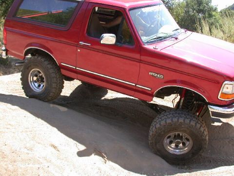 1996 Ford Bronco - Close passenger-side view of a red 1992-96 Ford Bronco articulating on a dirt mound, with the front wheel dropped and the red coil-over sho…