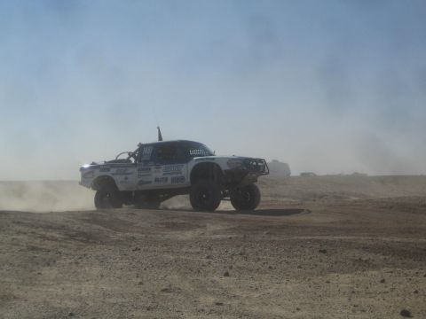 1990 Ford Bronco - Photograph of a white off-road race truck covered in sponsor decals racing in the desert, not a technical document.