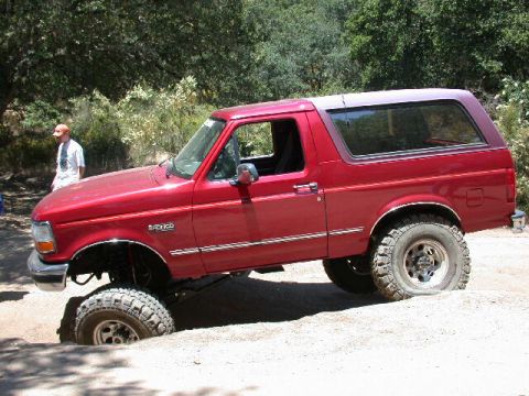1996 Ford Bronco - Full driver-side profile of a red 1992-96 Ford Bronco on a dirt trail, front wheel dropped into a low spot so the suspension articulates. V…