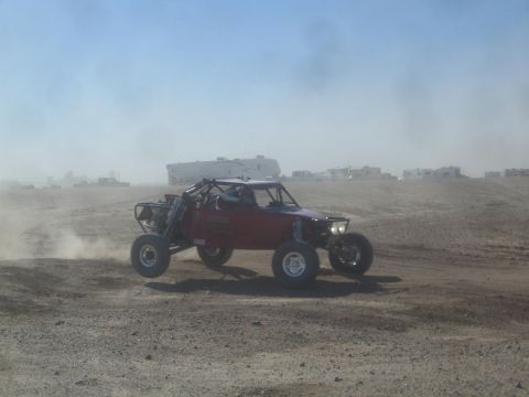 1990 Ford Bronco - Photograph of a red off-road race buggy racing past RVs in a dusty desert, not a technical document.