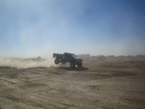 1990 Ford Bronco - Photograph of a dark off-road race truck jumping in a dusty desert, not a technical document.