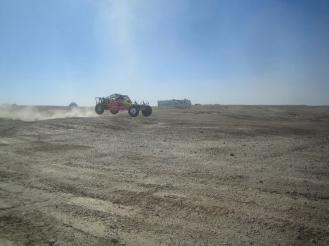 1990 Ford Bronco - Action photo of a red tube-frame off-road sand car kicking up dust on a desert race course, not a technical document.