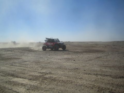 1990 Ford Bronco - Action photo of a red DEW Racing off-road race car driving through dust, not a technical document.
