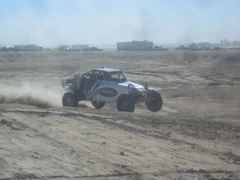 1990 Ford Bronco - Action photo of a white and blue off-road race car drifting through dust on a desert track, not a technical document.