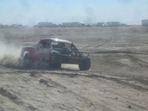 1990 Ford Bronco - Action photo of a red off-road race truck sliding through dust on a desert course, not a technical document.