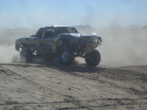 1990 Ford Bronco - Action photo of an off-road trophy truck racing through dust on a desert track, not a technical document.