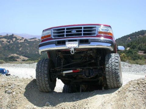 1996 Ford Bronco - A lifted red pickup truck navigates a dirt path with a blue vehicle visible in the distance.