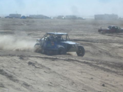 1990 Ford Bronco - Action photo of a blue off-road tube-frame buggy sliding through dust on a desert course, not a technical document.