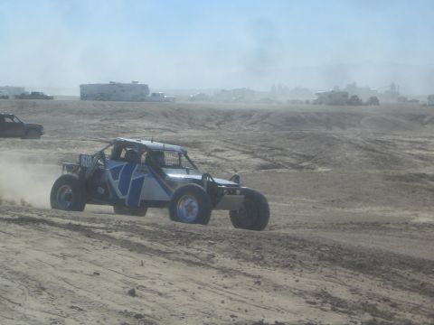1990 Ford Bronco - Action photo of a blue and white off-road buggy racing across a dusty desert track, not a technical document.