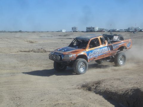 1990 Ford Bronco - Action photo of an orange off-road race truck number 700 racing through dust, not a technical document.