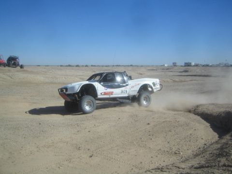 1990 Ford Bronco - Action photo of a white off-road race truck number 1479 racing across a dusty desert track, not a technical document.