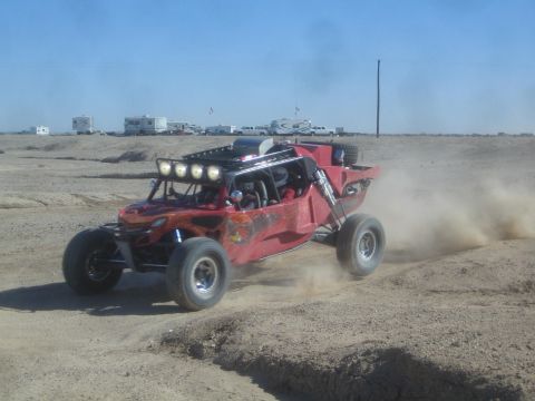 1990 Ford Bronco - Action photo of a red off-road buggy with a roof light bar racing through dust on a desert course, not a technical document.