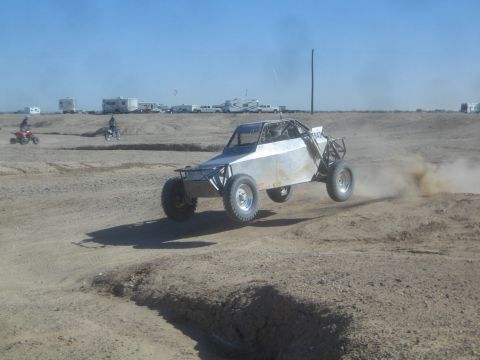 1990 Ford Bronco - Action photo of a silver tube-frame off-road buggy racing across a dusty desert track, not a technical document.