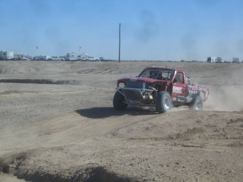 1990 Ford Bronco - Action photo of a red off-road race truck racing through dust on a desert track, not a technical document.