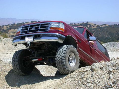 1996 Ford Bronco - Low front three-quarter shot of a red 1992-96 Ford Bronco perched on a rocky rise, front driver-side wheel drooping to reveal the solid fro…