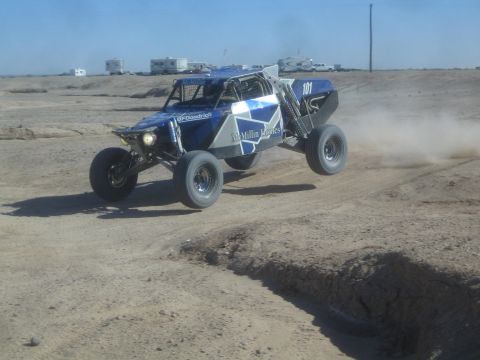 1990 Ford Bronco - Action photo of a blue and white off-road buggy number 101 racing across a dusty desert track, not a technical document.