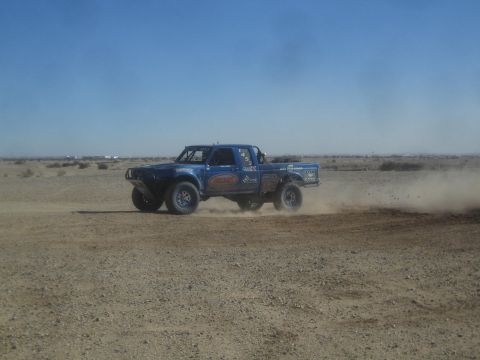 1990 Ford Bronco - Action photo of a blue off-road race truck racing through dust on a desert course, not a technical document.