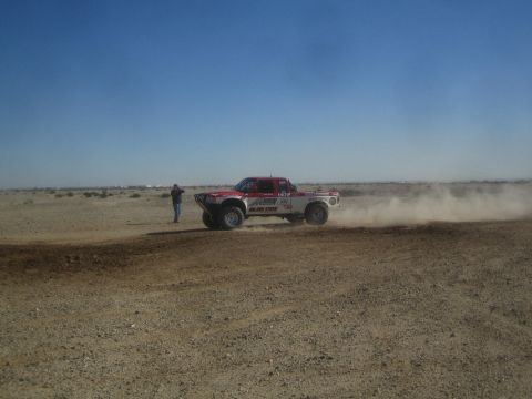 1990 Ford Bronco - Action photo of a red and white off-road race truck racing past a bystander on a dusty desert track, not a technical document.