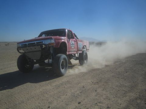 1990 Ford Bronco - Action photo of a red off-road race truck (number 1487) kicking up dust on a desert course; not a technical document.