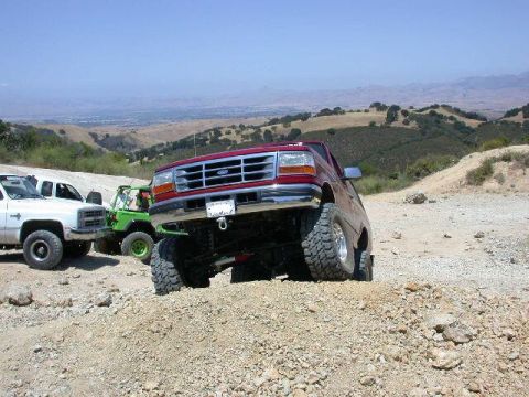 1996 Ford Bronco - A red 1990s full-size Ford Bronco tipped up on a gravel mound at a hilltop off-road park, seen from the low front three-quarter with its li…