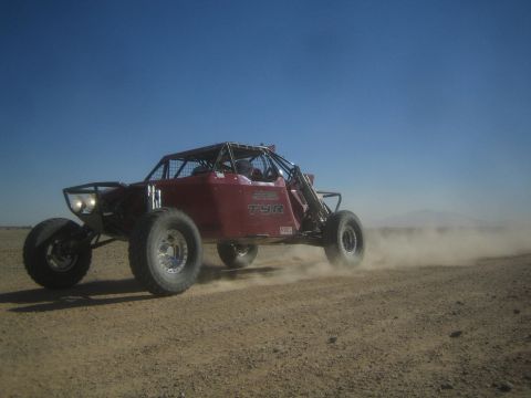 1990 Ford Bronco - Action photo of a maroon off-road buggy with lights on racing across a desert course; not a technical document.