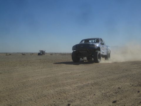 1990 Ford Bronco - Action photo of a blue off-road race truck on a desert course with a chase vehicle following; not a technical document.