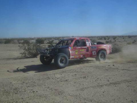 1990 Ford Bronco - Action photo of a red sponsor-decaled off-road race truck on a desert course; not a technical document.