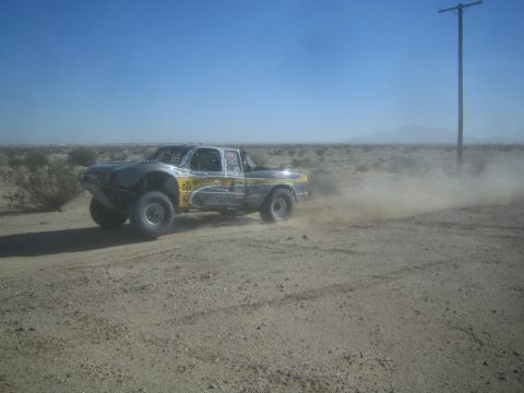 1990 Ford Bronco - Action photo of a silver and yellow off-road race truck driving along a dusty desert road; not a technical document.