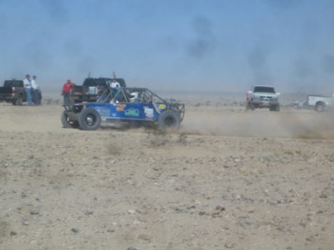 1990 Ford Bronco - Action photo of a blue off-road buggy racing past spectators and chase trucks in the desert; not a technical document.