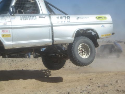 1990 Ford Bronco - Close action photo of a silver off-road race truck (number 1478) catching air over a desert jump; not a technical document.